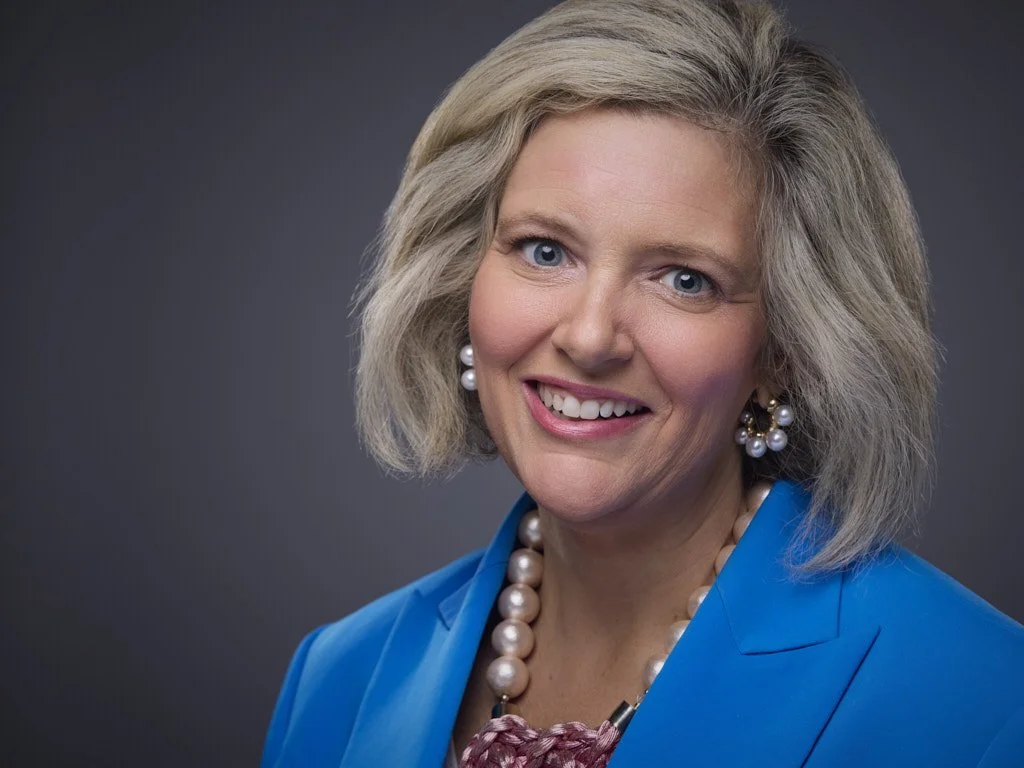 Professional headshot of a middle-aged woman with blonde hair, wearing a blue blazer, pearl earrings, and a pearl necklace, smiling against a gray background.