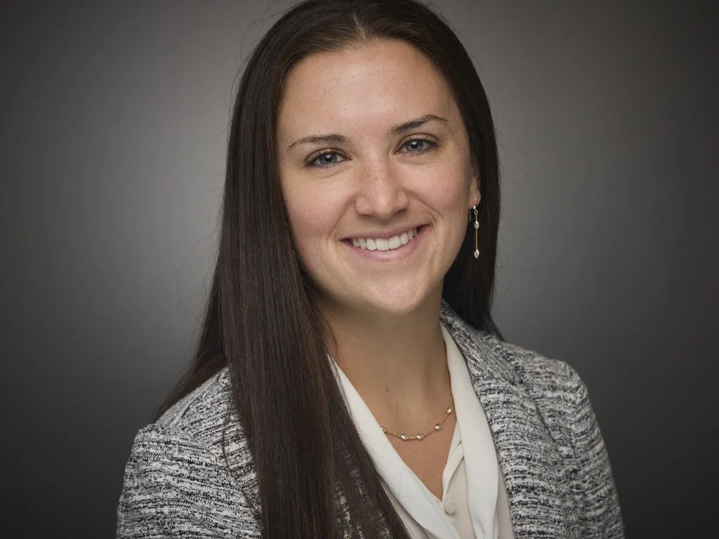 Headshot of a woman with long brown hair, smiling, wearing a gray patterned blazer, white blouse, and gold jewelry, posed against a gray background.