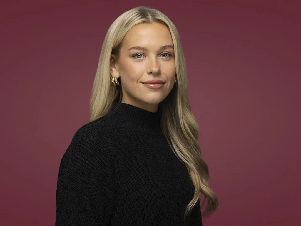 Professional headshot of a woman with long blonde hair wearing a black turtleneck, standing against a maroon background.