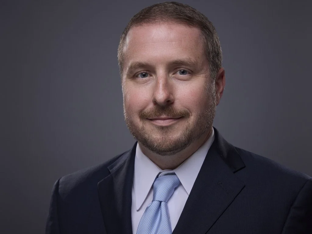 Professional headshot of a man with short light brown hair, blue eyes, wearing a dark suit, light blue tie, and white shirt, smiling against a gray background.