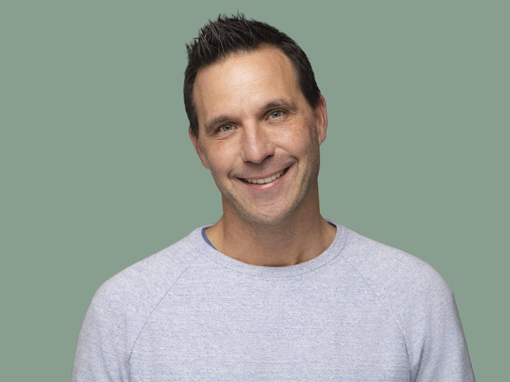 A smiling man with short dark hair wearing a light gray crewneck shirt against a muted green background.
