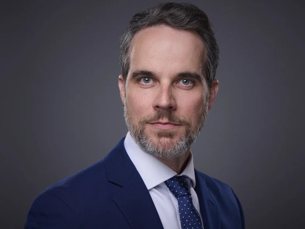 A professional headshot of a middle-aged man with short dark hair and a beard, wearing a navy suit, white shirt, and a navy tie with white polka dots, against a gray background.
