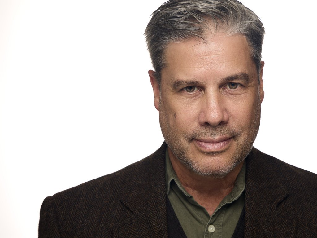 Close-up portrait of a middle-aged man with gray hair, beard, wearing a dark brown blazer and green shirt, against a plain white background.