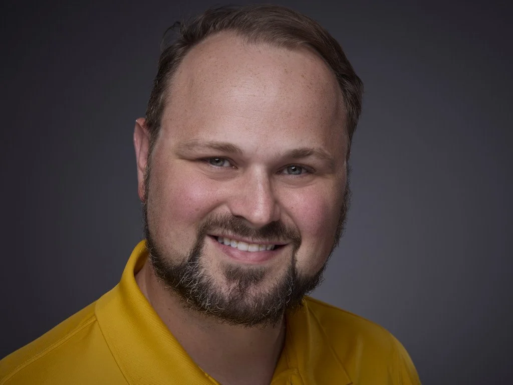 Professional headshot of a smiling man with short hair and a beard, wearing a yellow shirt, against a dark gray background.