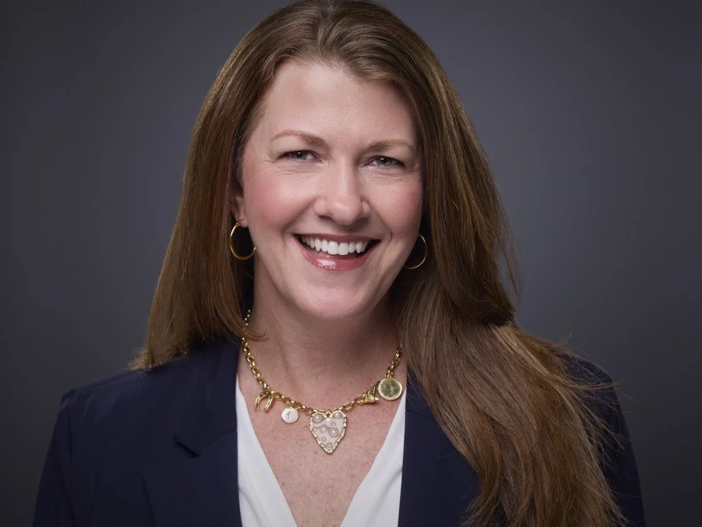 Headshot of a smiling woman with long red hair, wearing a navy blazer, jewelry, and a white top against a dark background.