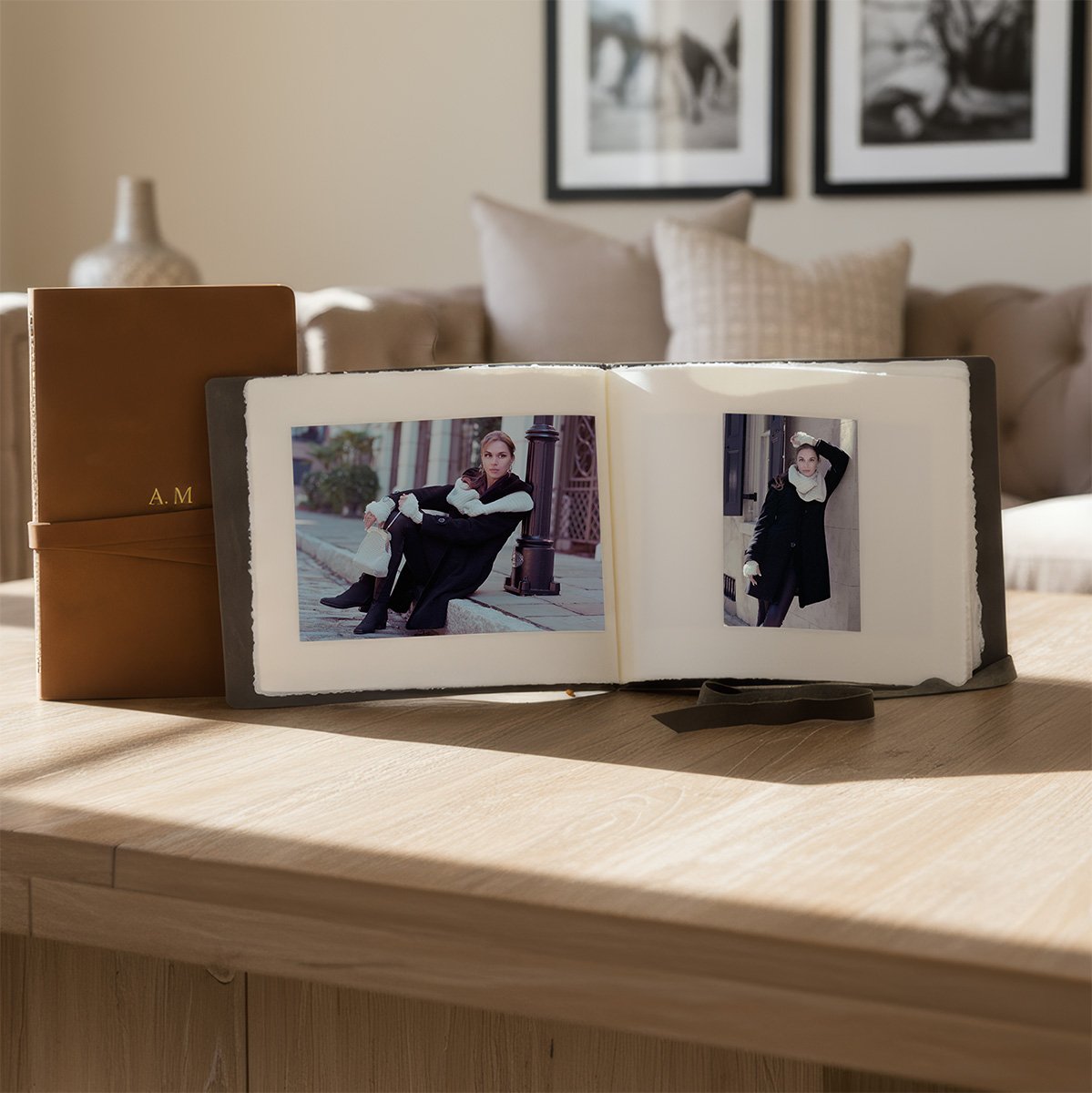 Open photo album with two pictures of a woman in black coat and gray scarf, sitting on a sidewalk and standing by a building. The album is on a wooden table in a living room with a beige sofa and framed black-and-white photos on the wall in the background.