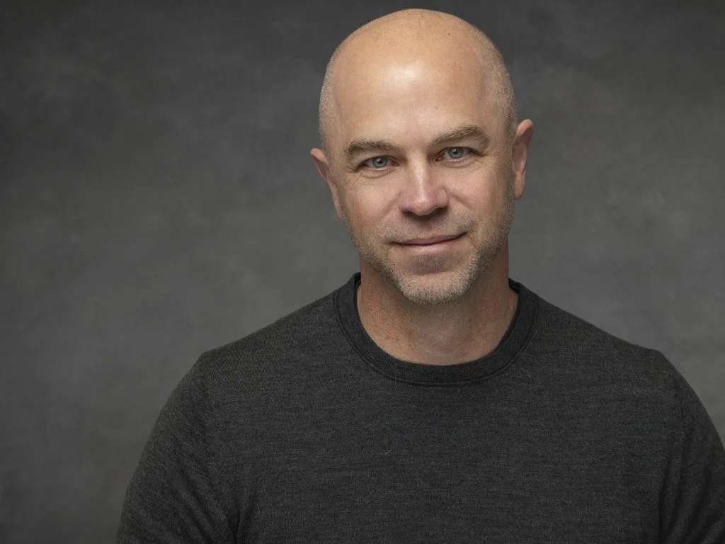 Headshot of a bald man with light skin, wearing a black shirt, against a dark gray background.