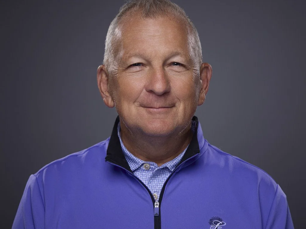 Professional headshot of a middle-aged man with short gray hair and a slight smile, wearing a blue quarter-zip jacket over a collared shirt, standing against a dark gray background.