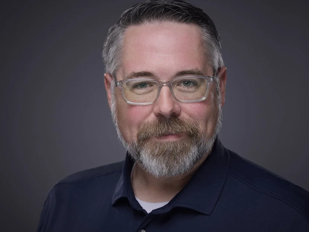 Professional headshot of a middle-aged man with gray hair and beard wearing glasses and a dark collared shirt, posing against a dark gray background.