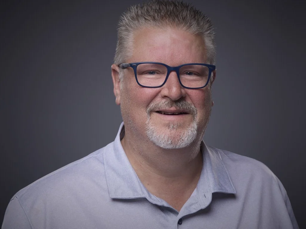Professional headshot of a middle-aged man with light skin, gray hair, and a beard, wearing glasses with dark blue frames and a light gray collared shirt, smiling against a dark gray background.