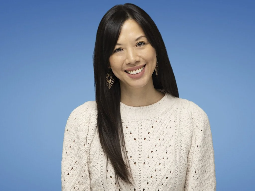 A young woman with long dark hair smiling, wearing a cream knit sweater and earrings, against a blue background.