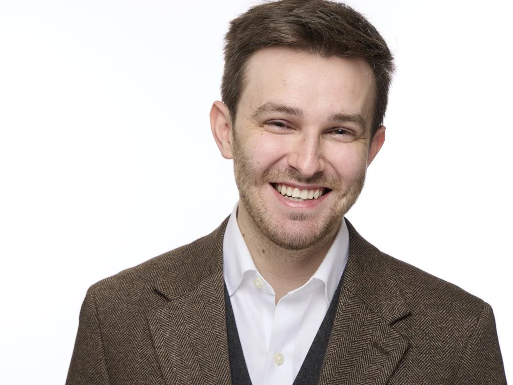 Portrait of a smiling man with brown hair, wearing a white shirt and brown blazer, against a plain white background.