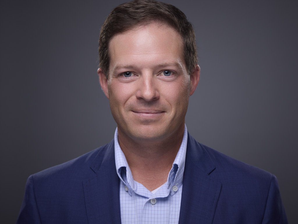 Professional headshot of a man wearing a blue blazer and light blue collared shirt against a dark gray background.