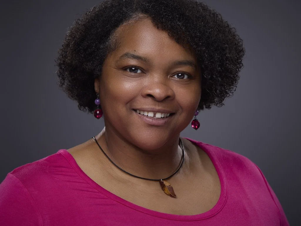 Headshot of a woman wearing a pink top, purple and red earrings, a black choker, and a necklace with a pendant, smiling against a dark gray background.