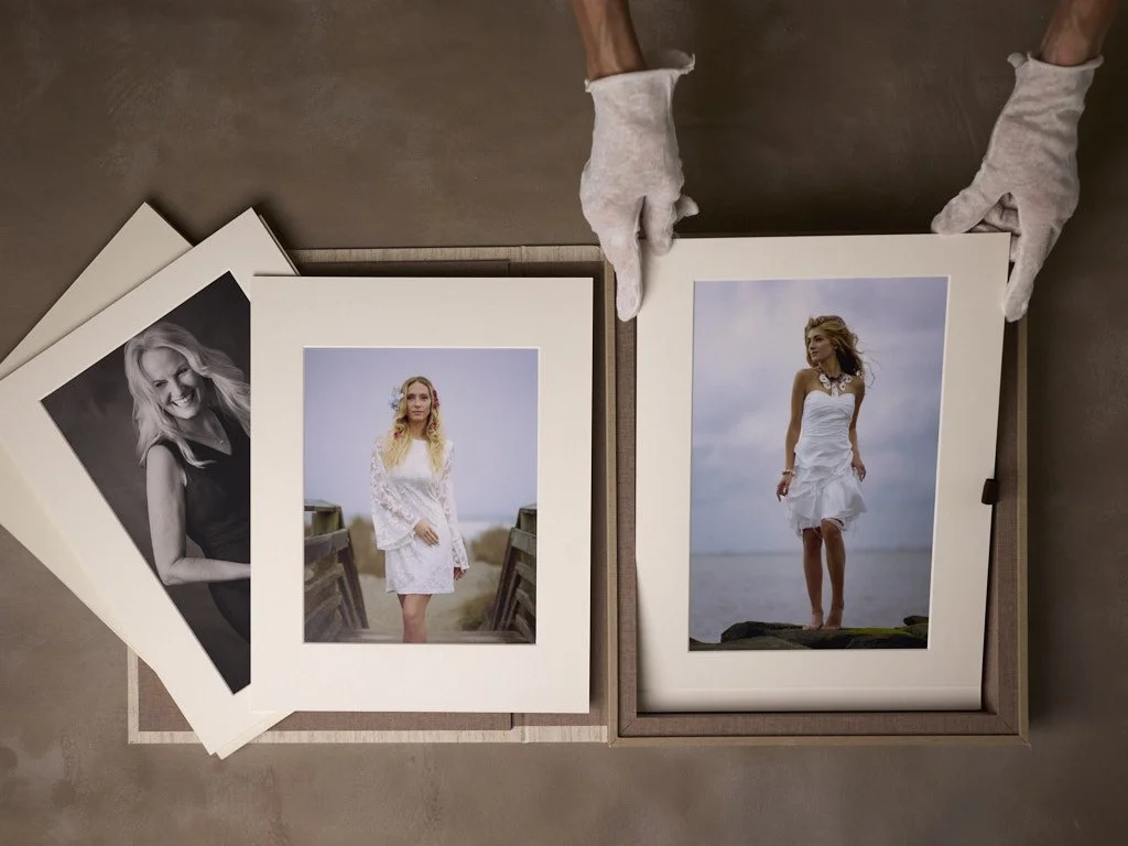 A person wearing white gloves handling matted photographs of women in white dresses, with two additional black-and-white photos beside a colored one showing a woman with long blonde hair.