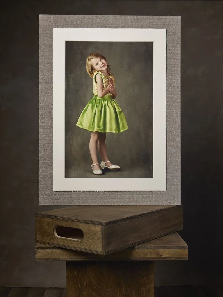 A framed photograph of a young girl in a green dress, smiling and standing against a neutral background, placed on a wooden stand.