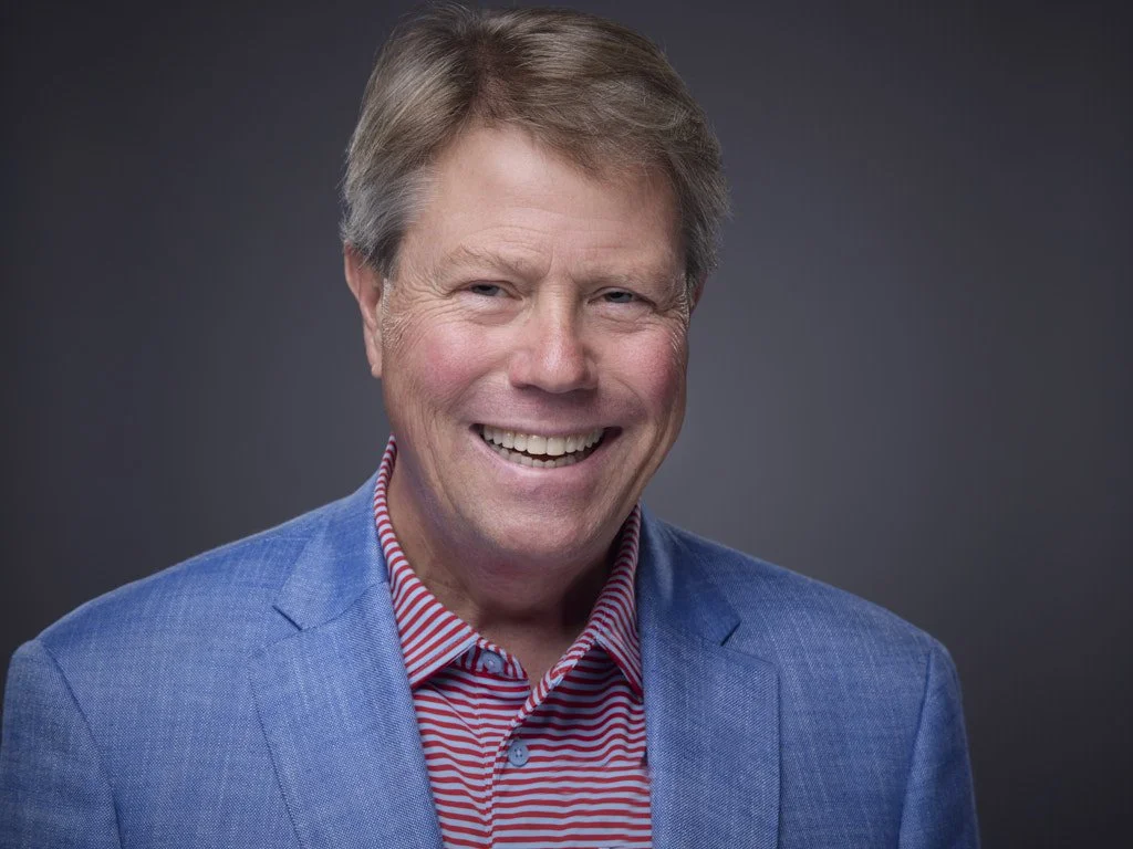 Professional headshot of a smiling man with light brown hair, wearing a blue blazer and a red and white striped shirt, against a dark gray background.