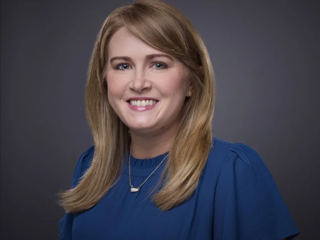 Professional headshot of a woman with blonde hair wearing a blue top and a gold necklace, smiling at the camera against a dark gray background.