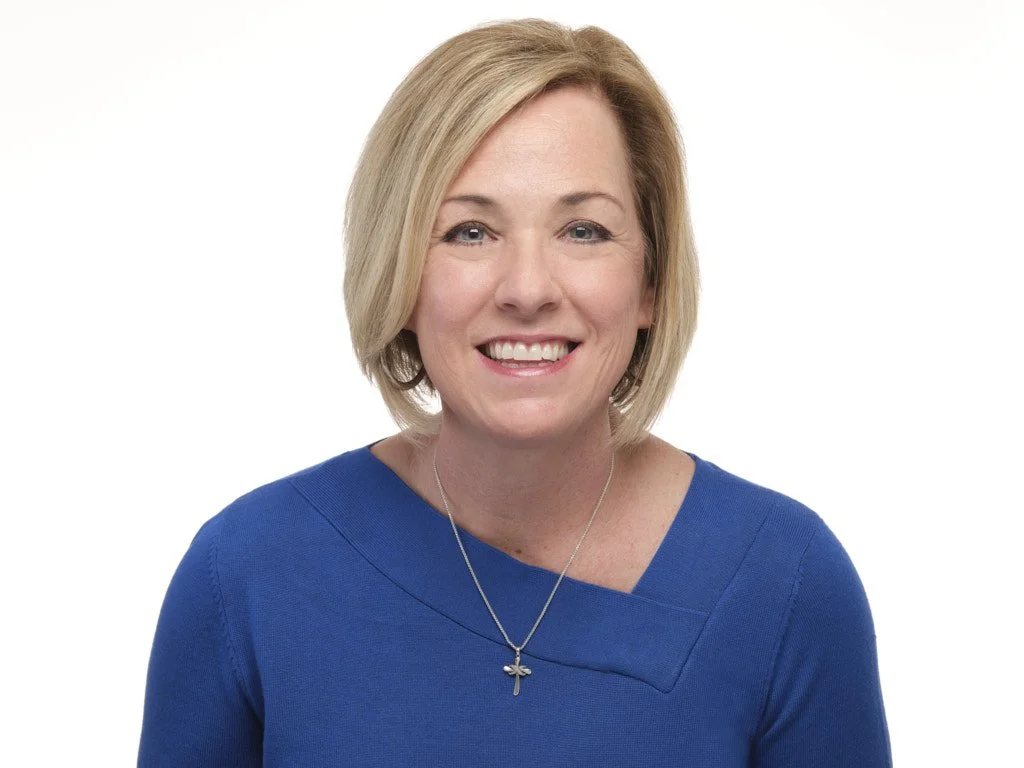 Professional headshot of a smiling woman with short blonde hair wearing a blue top and a silver necklace with a cross pendant