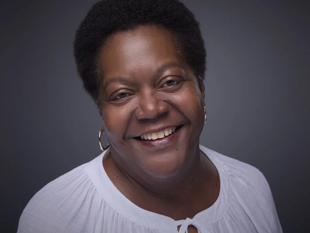 Professional headshot of a smiling Black woman with short natural hair, wearing hoop earrings and a white blouse against a dark gray background.