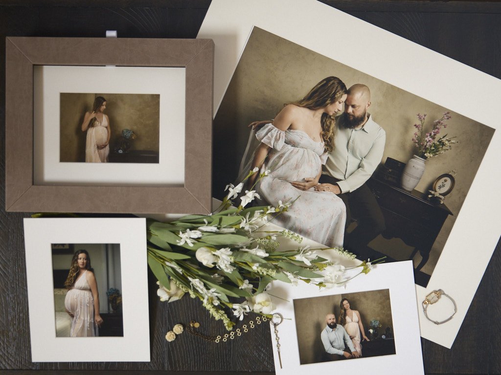 Collection of printed maternity photographs featuring a pregnant woman and her partner, a bouquet of white flowers, and jewelry on a dark wooden surface.