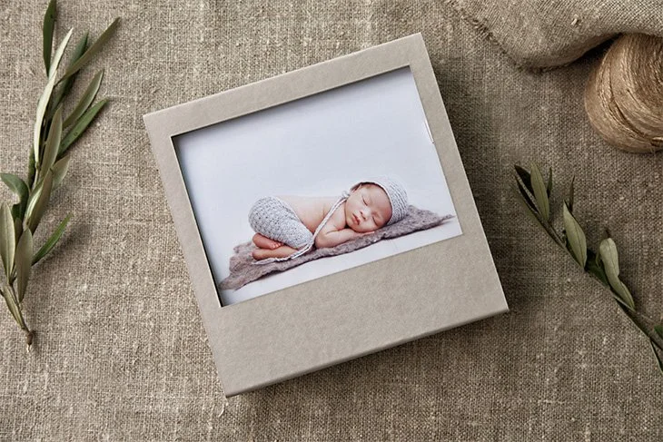 A framed photo of a sleeping baby in a gray knitted outfit and hat, lying on a soft gray surface, placed on a textured beige fabric background with sprigs of greenery around it.