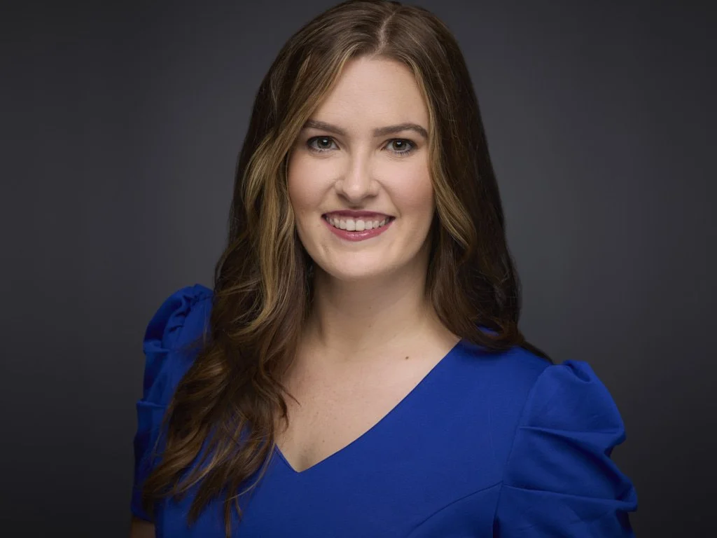 A portrait of a young woman with long brown hair, wearing a blue top, smiling against a dark gray background.