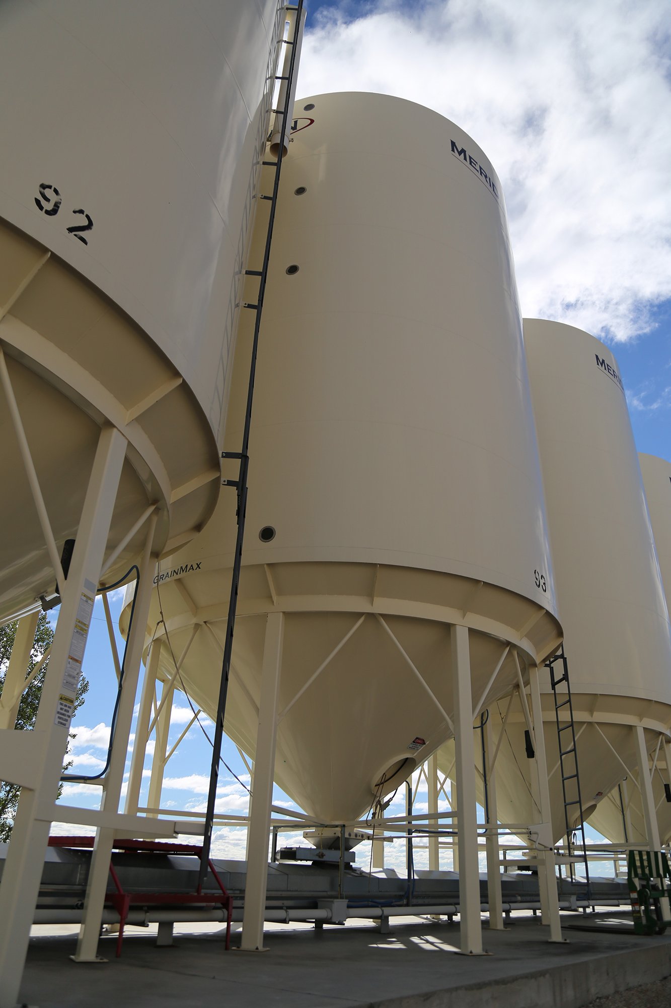 Large white storage tanks with black railings and ladders, set outdoors under a partly cloudy sky.