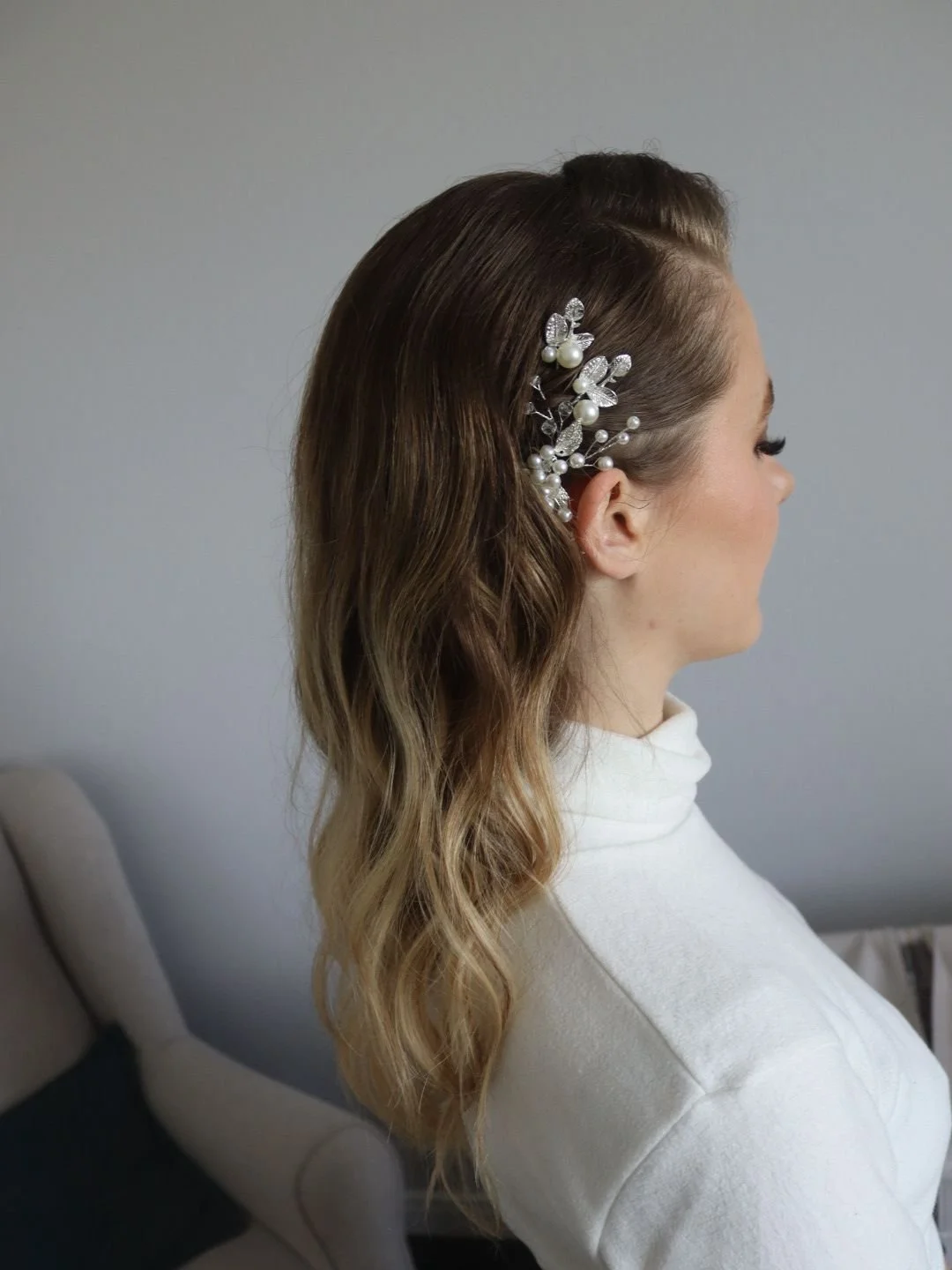 Une femme vue de profil avec des cheveux longs et ondulés, portant une barrette ornée de perles et de feuilles brillantes à l'oreille, vêtue d'un haut blanc.