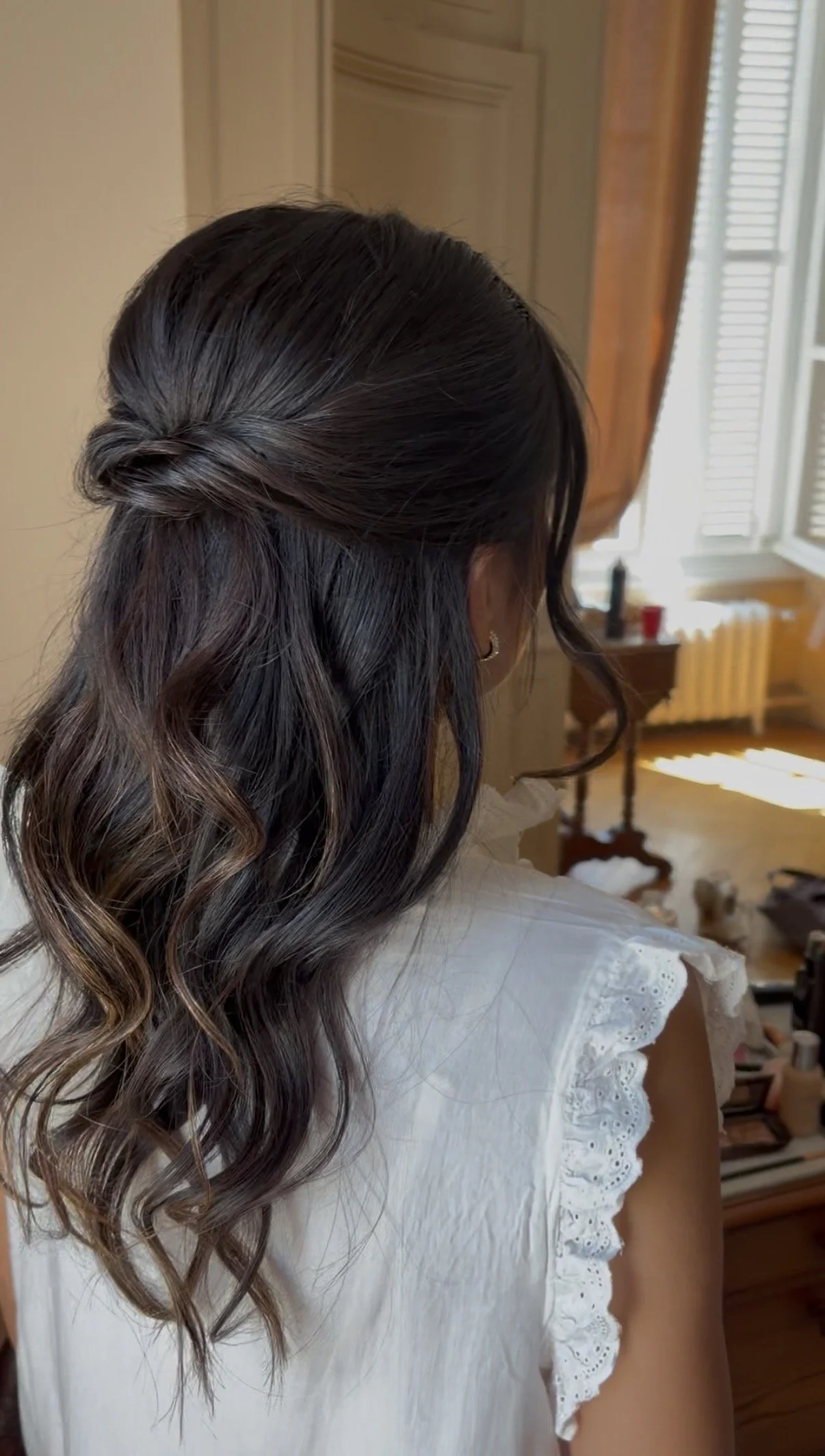 Une femme avec des cheveux mi longs, ondulés, dans un intérieur lumineux, portant une chemise blanche à manches courtes avec des détails en dentelle.