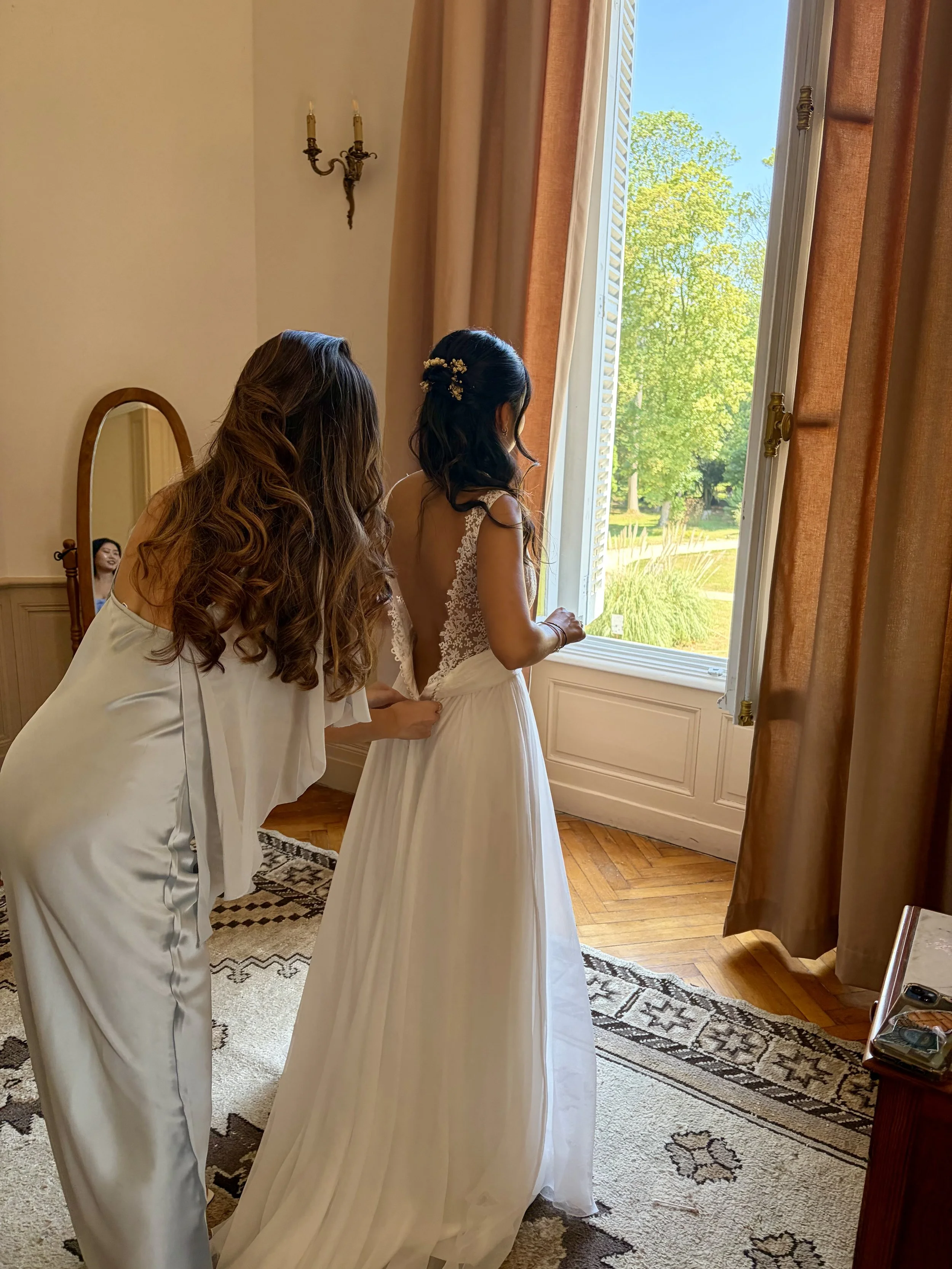 Deux femmes en robe de mariage regardent par une grande fenêtre ouverte sur un jardin ensoleillé, l'une d'elles ajustant la robe de la mariée.