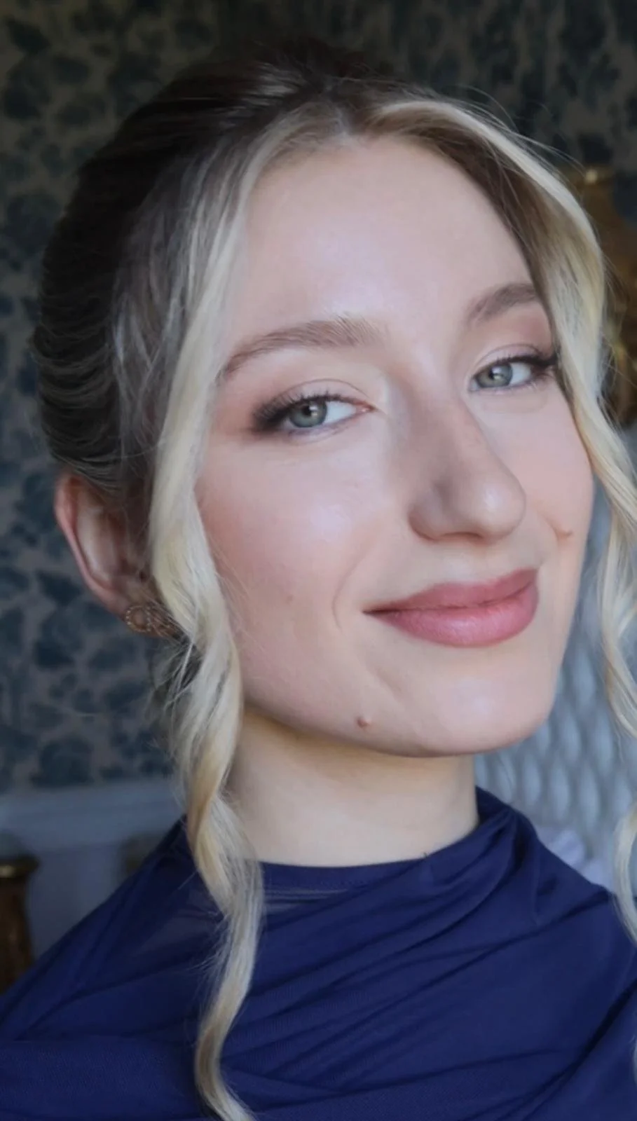 Jeune femme avec cheveux blonds ondulés, maquillage léger, portant une robe bleue, souriant légèrement dans un intérieur avec un mur sombre en arrière-plan.