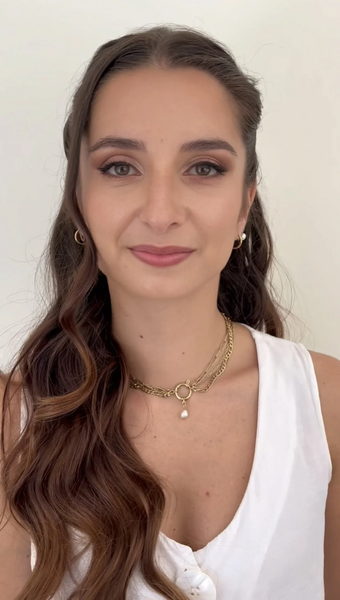 Portrait d'une jeune femme avec de longs cheveux bruns ondulés portant des bijoux en or et une chemise blanche.