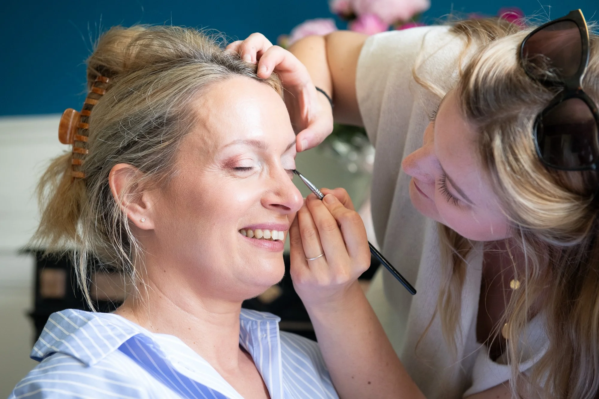 Une femme se fait maquiller par une artiste maquilleuse, qui applique du maquillage autour de ses yeux, dans un environnement intérieur.