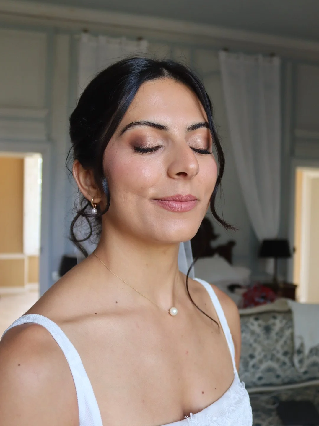 Femme avec des yeux fermés, maquillée, portant des bijoux et un haut blanc, dans une pièce lumineuse.