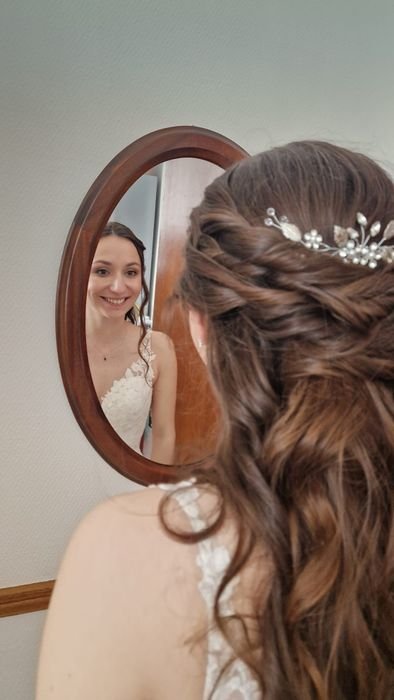 Une femme avec des cheveux bouclés portant une couronne de fleurs regarde son reflet dans un miroir oval, portant une robe de mariée