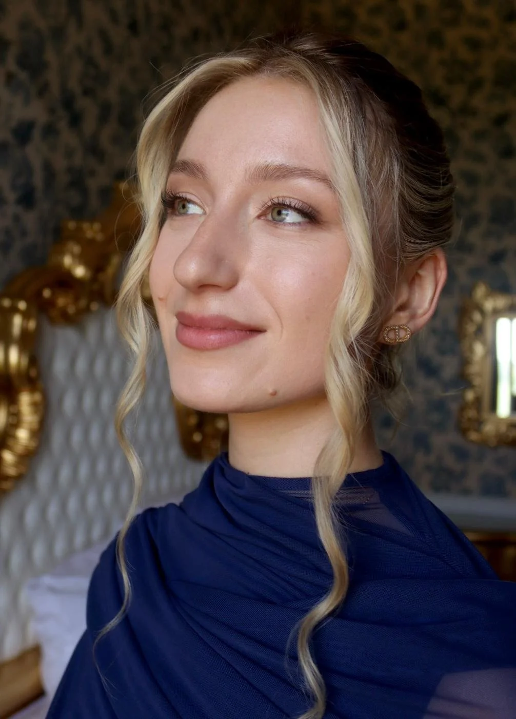 Une femme avec des cheveux blondes bouclés, portant des boucles d'oreilles dorées et un vêtement bleu, dans une pièce élégante avec un miroir doré et un lit à tête rembourrée.