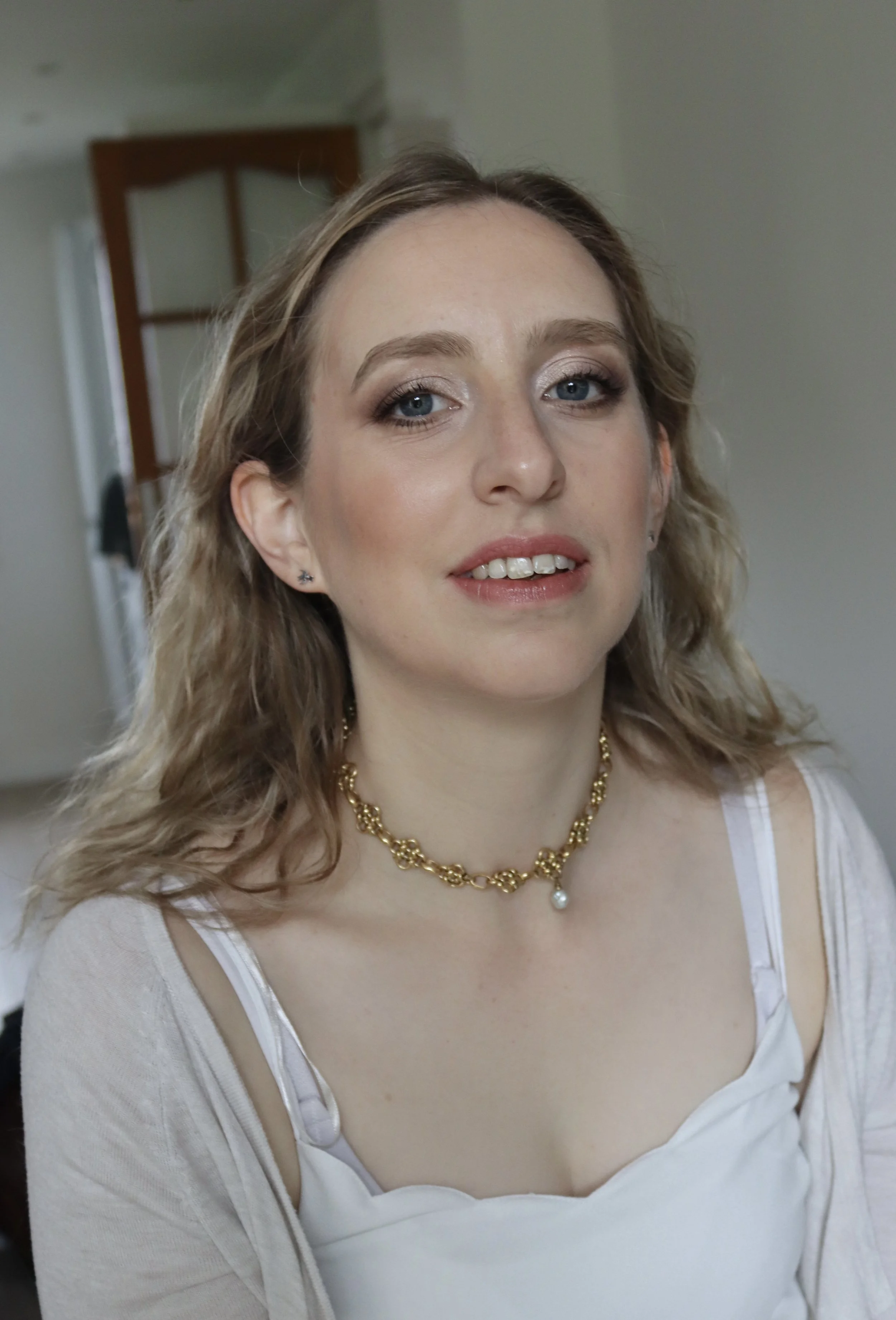 Portrait d'une femme avec des cheveux bruns ondulés, portant des yeux maquillés, un collier en or avec une perle, une robe blanche à bretelles, et un gilet beige, dans un intérieur domestique.