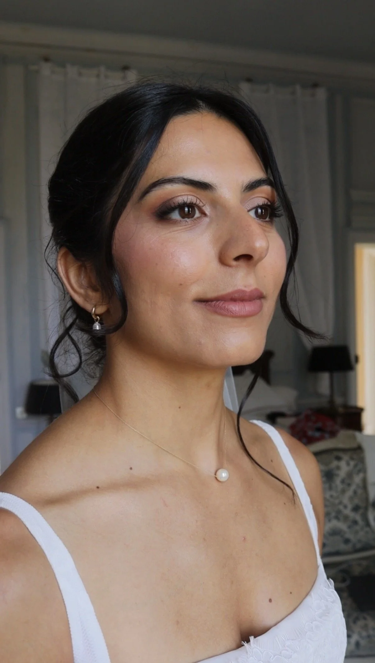 Jeune femme avec des cheveux noirs, maquillage sophistiqué, portant une robe blanche avec des bretelles, porte des boucles d'oreilles en perles et un collier.