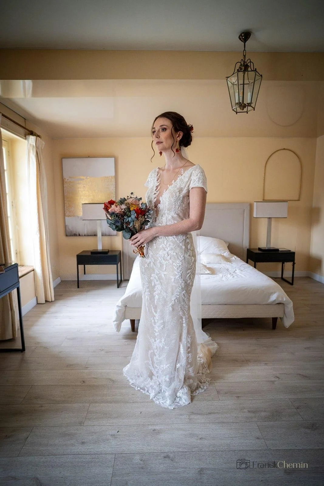 Une femme en robe de mariée blanche tient un bouquet de fleurs dans une chambre lumineuse