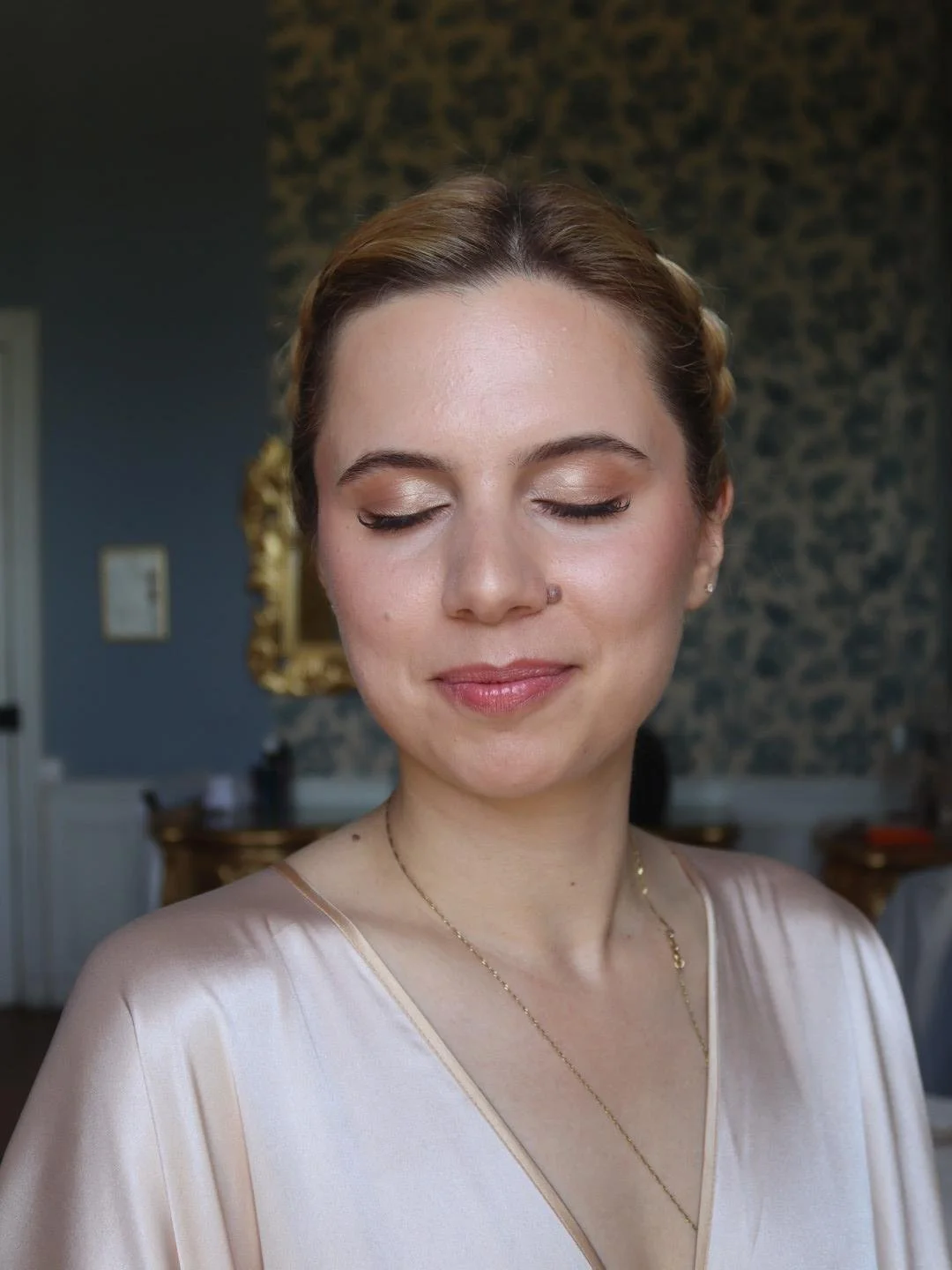 Une femme avec les yeux fermés, portant une robe en satin crème et un collier délicat, dans une pièce élégante avec un mur en papier peint animal print et un miroir doré en arrière-plan.