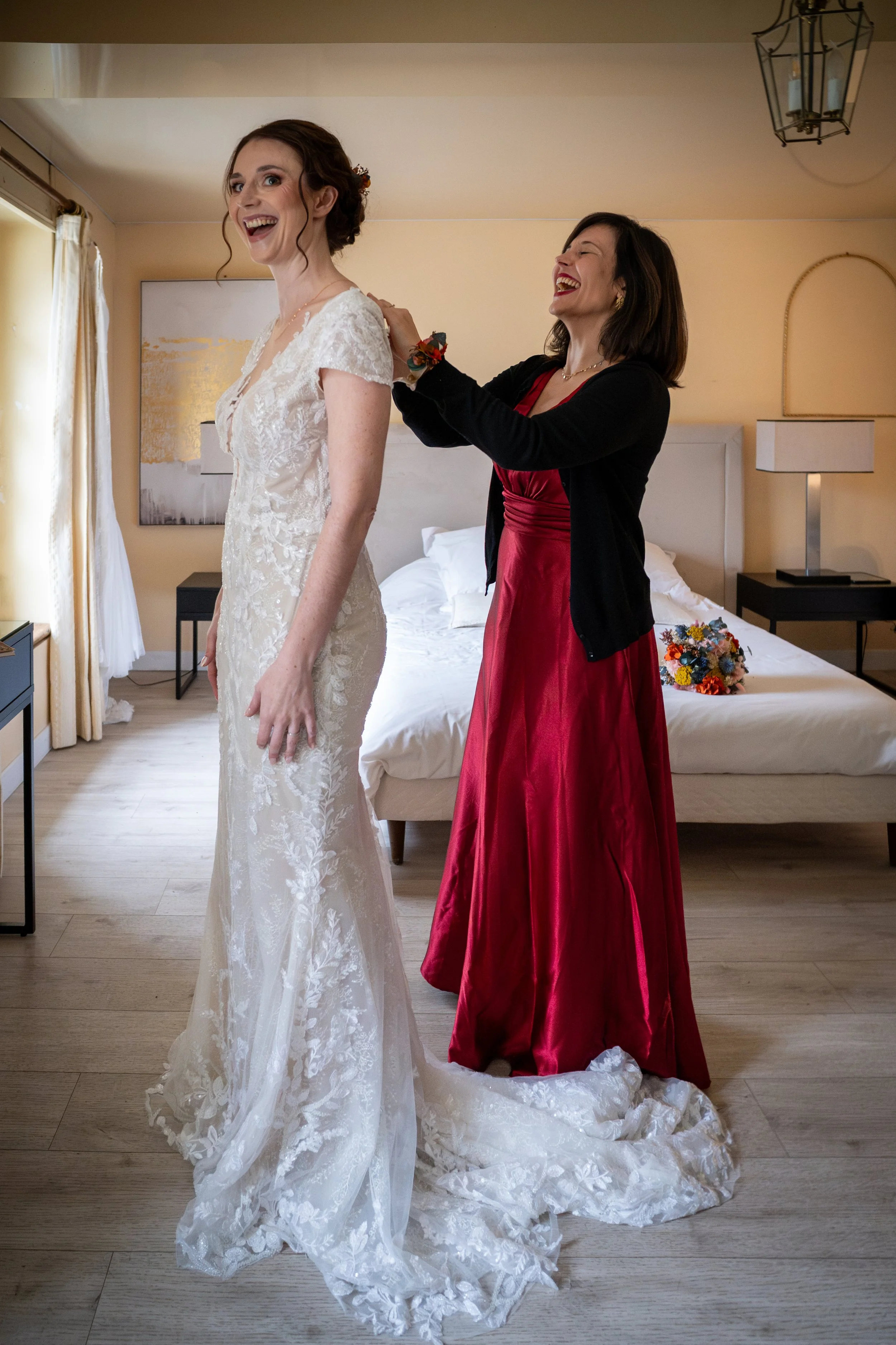 Une femme portant une robe de mariée blanche en dentelle avec un décolleté en V, et une autre femme en robe rouge avec une veste noire, qui l'aide à ajuster sa robe, dans une chambre avec un lit et des lampes de chevet.