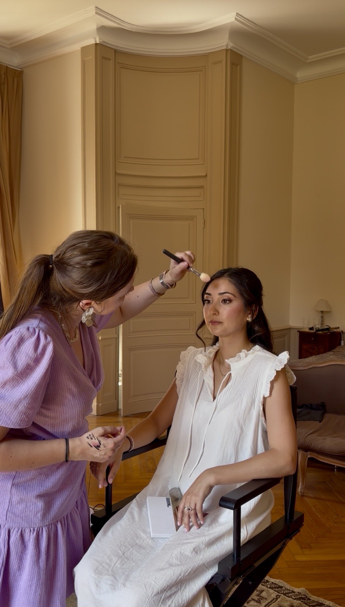 Une femme aux cheveux foncés et habillée en blanc est assise dans une chaise, une autre femme maquille ses lèvres avec un pinceau dans une pièce élégante avec des murs beige, des rideaux dorés et un mobilier ancien.