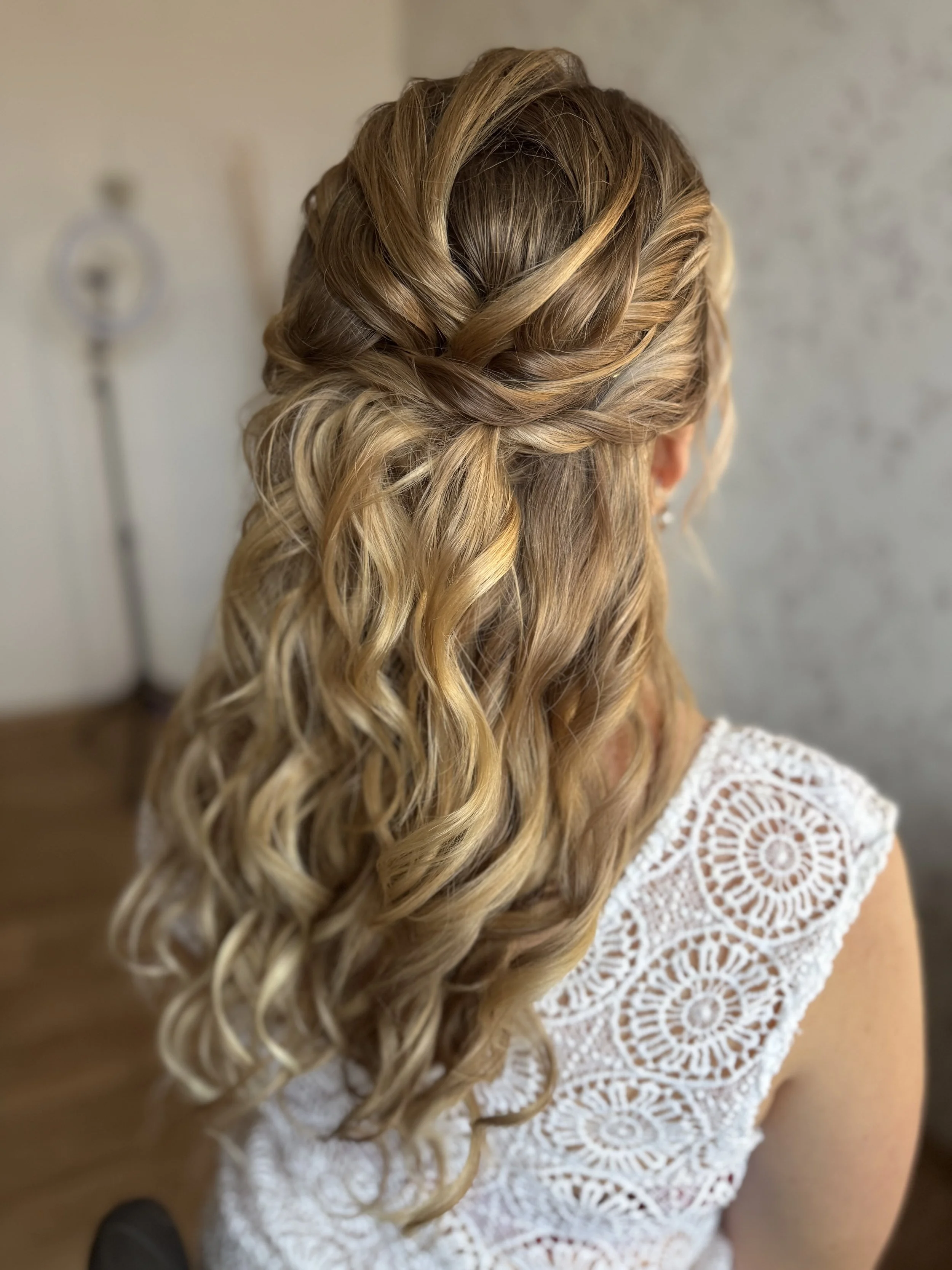 Une femme avec des cheveux longs et bouclés, coiffés dans un style avec des torsades sur le dessus de la tête, portant une robe blanche en dentelle avec un motif circulaire.