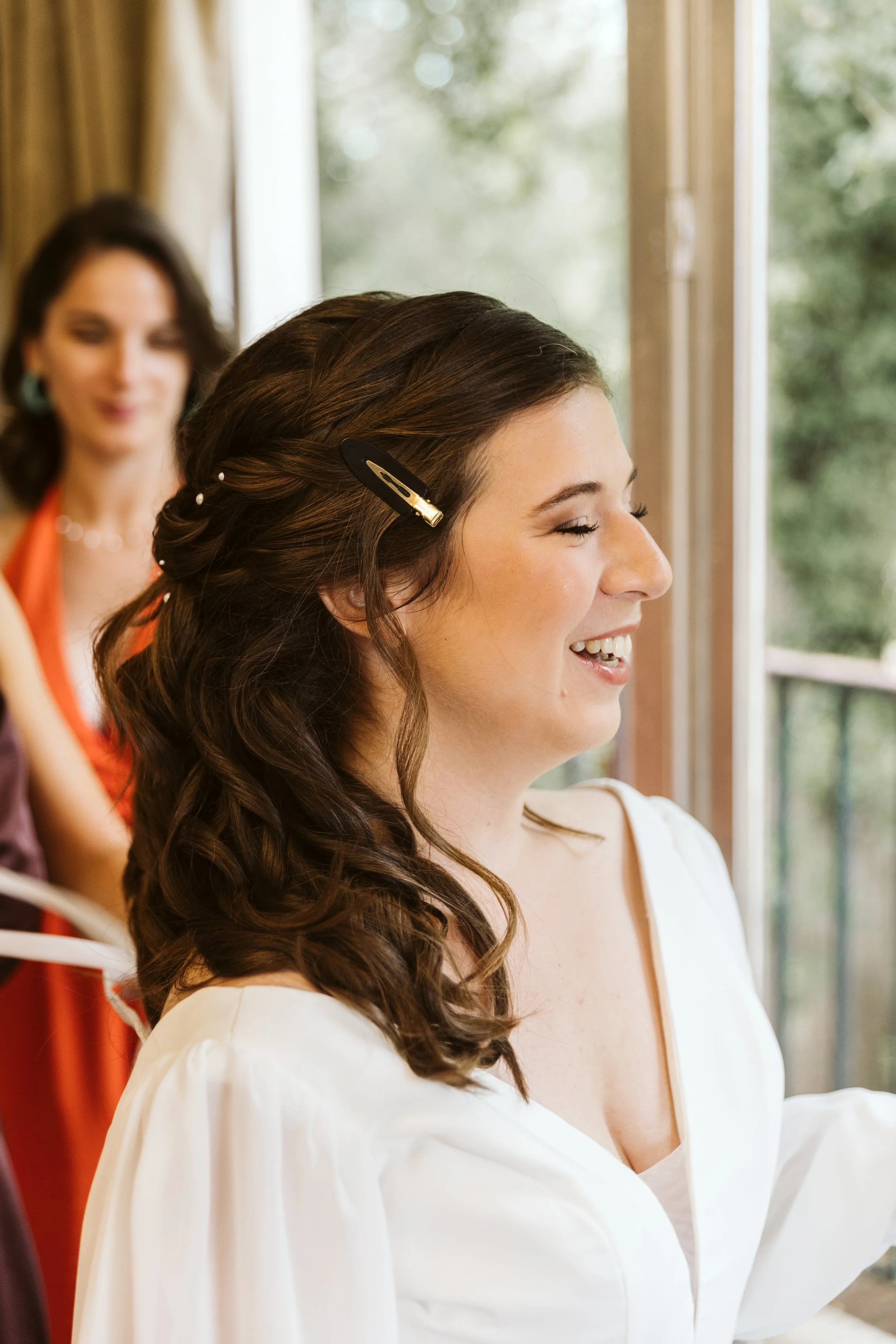Une femme souriante aux cheveux longs et bouclés, portant une chemise blanche, est en train de se coiffer avec des pinces dans les cheveux, avec un fond de fenêtre et une autre femme floue en arrière-plan.