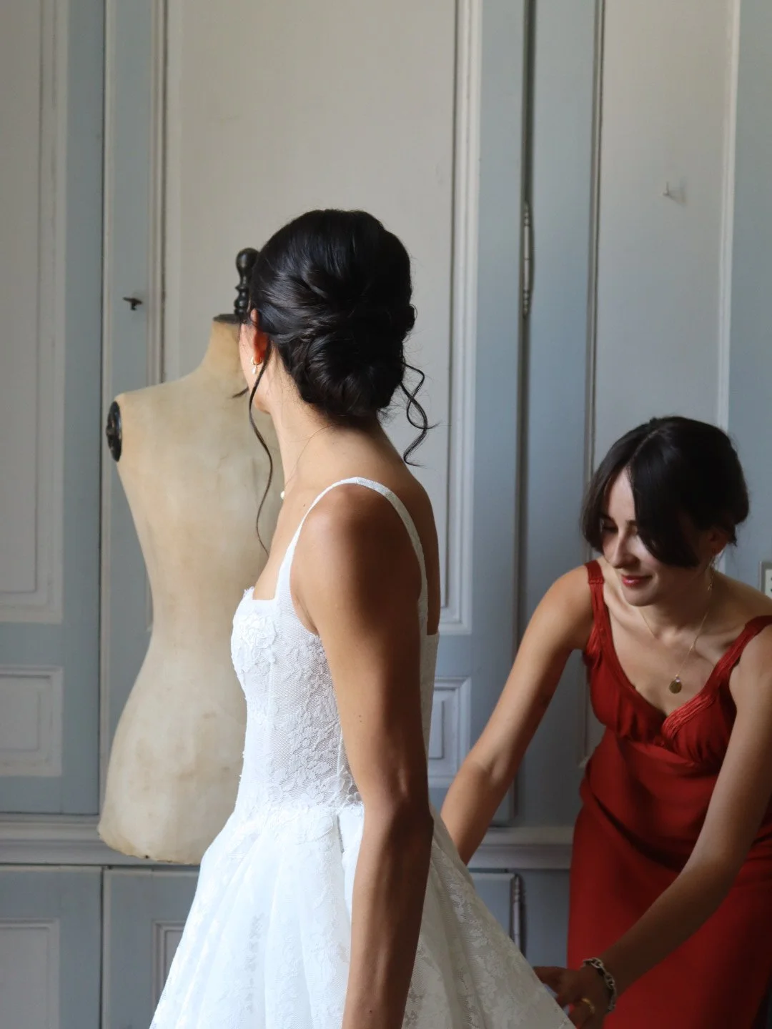 Une femme essaie une robe de mariée avec une femme qui l'aide. En arrière-plan, un mannequin et des placards de couleur pastel.