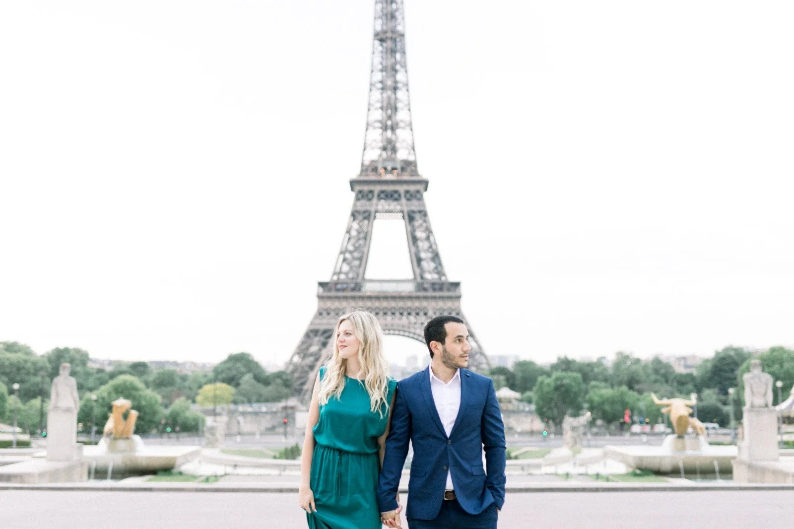 Un couple tenant la main devant la Tour Eiffel à Paris, la femme porte une robe verte et l'homme un costume bleu.