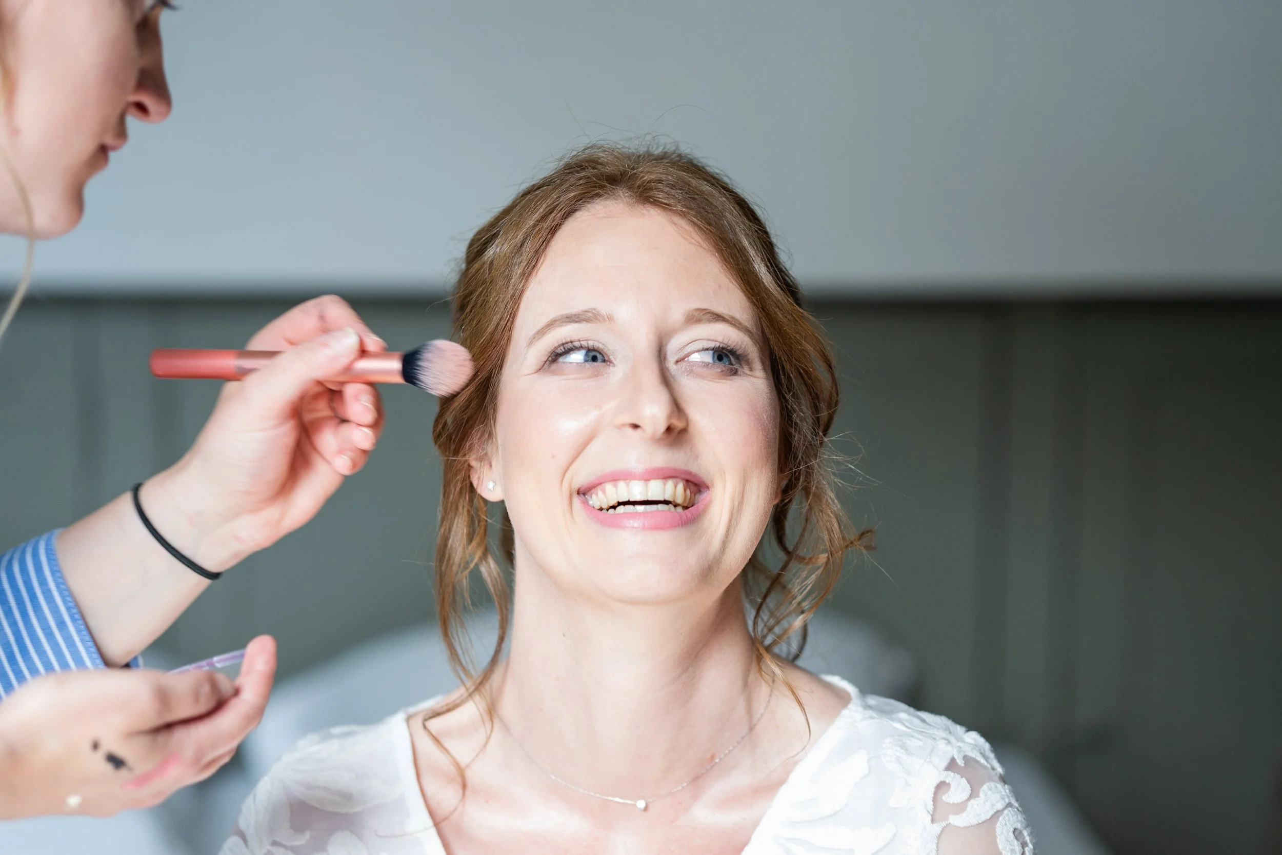 Une femme souriante reçoit une séance de maquillage, une main tient un pinceau près de son visage.