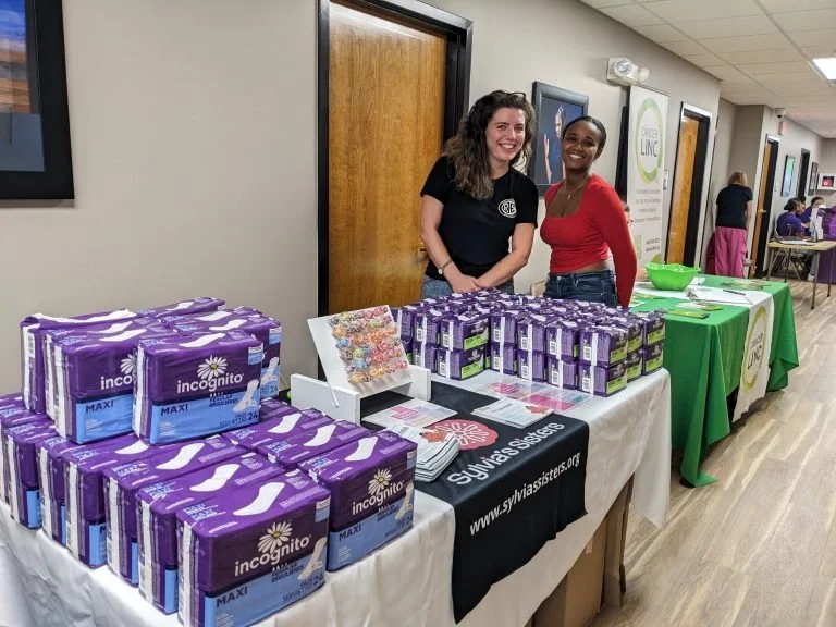 Two women standing behind a table with purple boxes of IncoGinto Maxi menstrual pads and other papers at an indoor event.