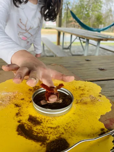 Child's hand showing off a brown pipe cleaner that has been made into a ring. The child's hand hovers above a yellow paper spread on a wooden table outdoors.